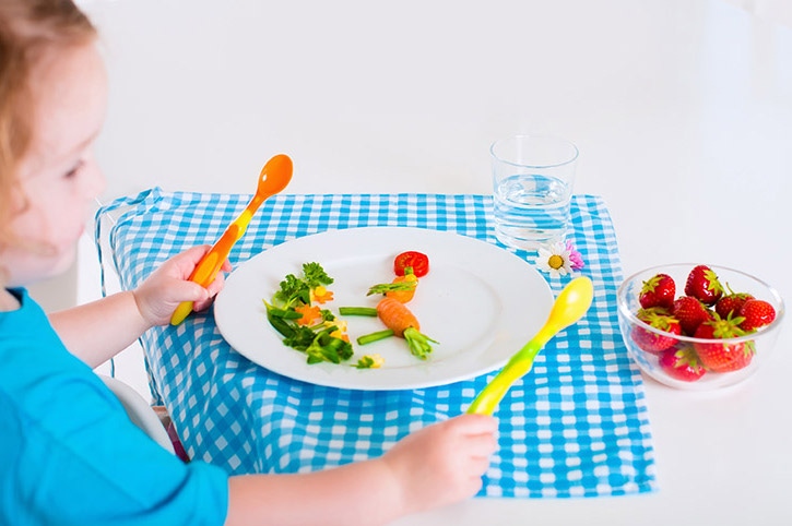 Bebe con cucharas en las dos manos y un platillo de comida con verduras para crear nuevos hábitos saludables